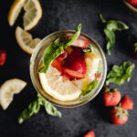 A glass filled with water, sliced lemon, strawberries, and basil leaves on a dark background, accompanied by additional lemon slices, strawberries, and basil.