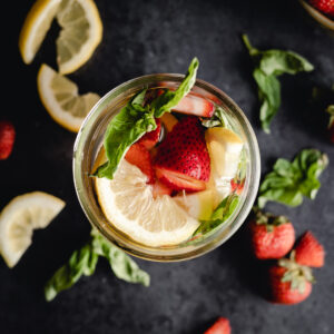 A glass filled with water, sliced lemon, strawberries, and basil leaves on a dark background, accompanied by additional lemon slices, strawberries, and basil.