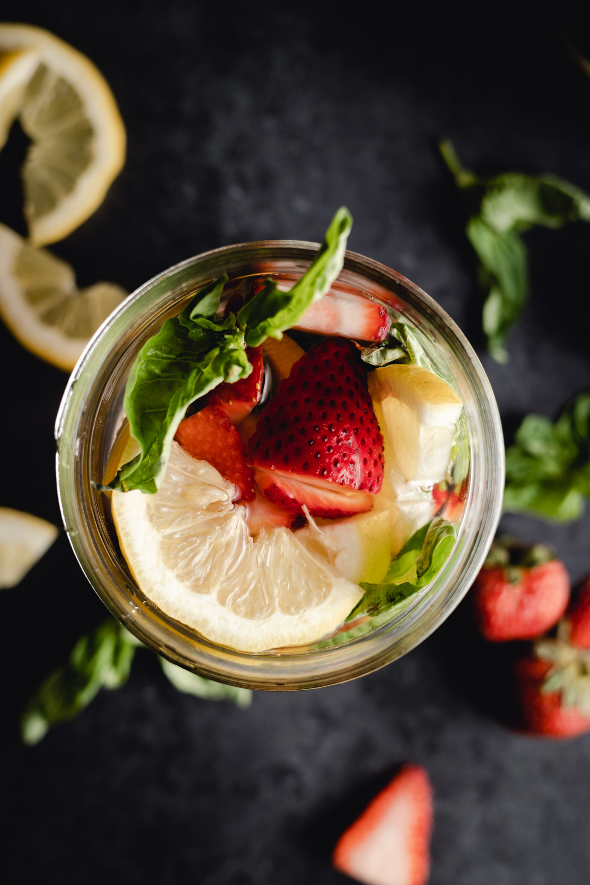 A top view of a glass filled with water, lemon slices, strawberries, mint leaves, and ice cubes. Fresh strawberries, lemon slices, and mint leaves are scattered around the glass on a dark surface.