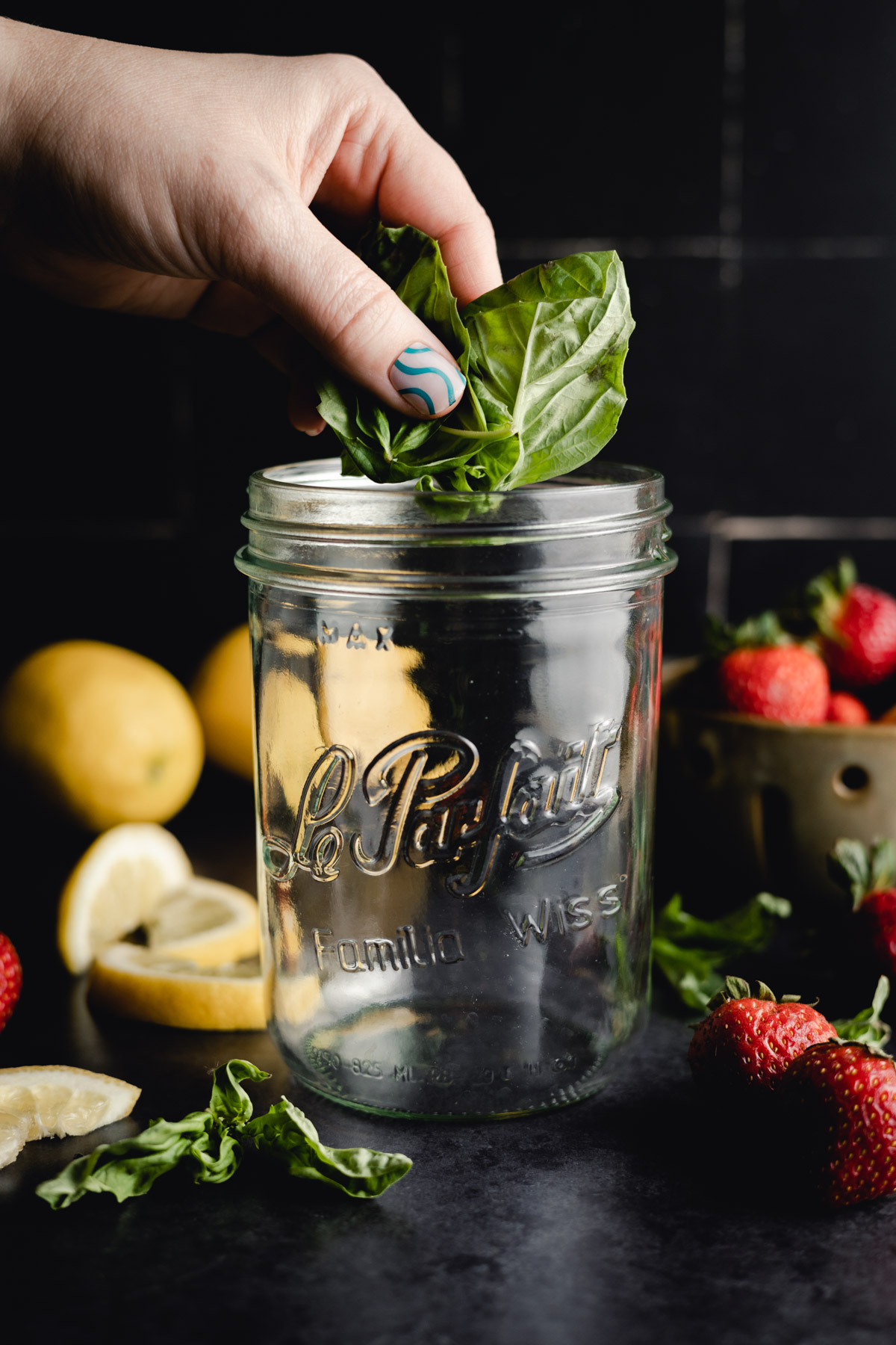 A hand places fresh basil leaves into a glass jar labeled "Le Parfait" with strawberries, lemons, and other basil leaves in the background.