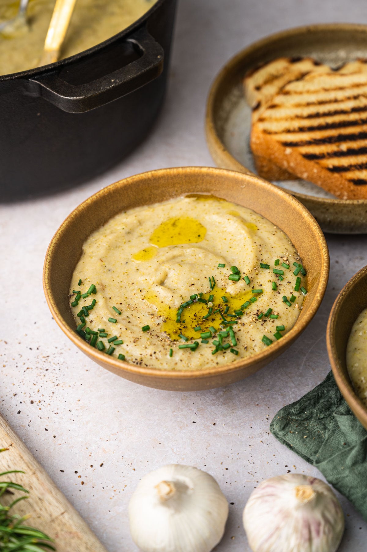 A bowl of creamy soup garnished with chives and olive oil, placed next to a plate of toasted bread. Garlic and a pot of soup are in the background.