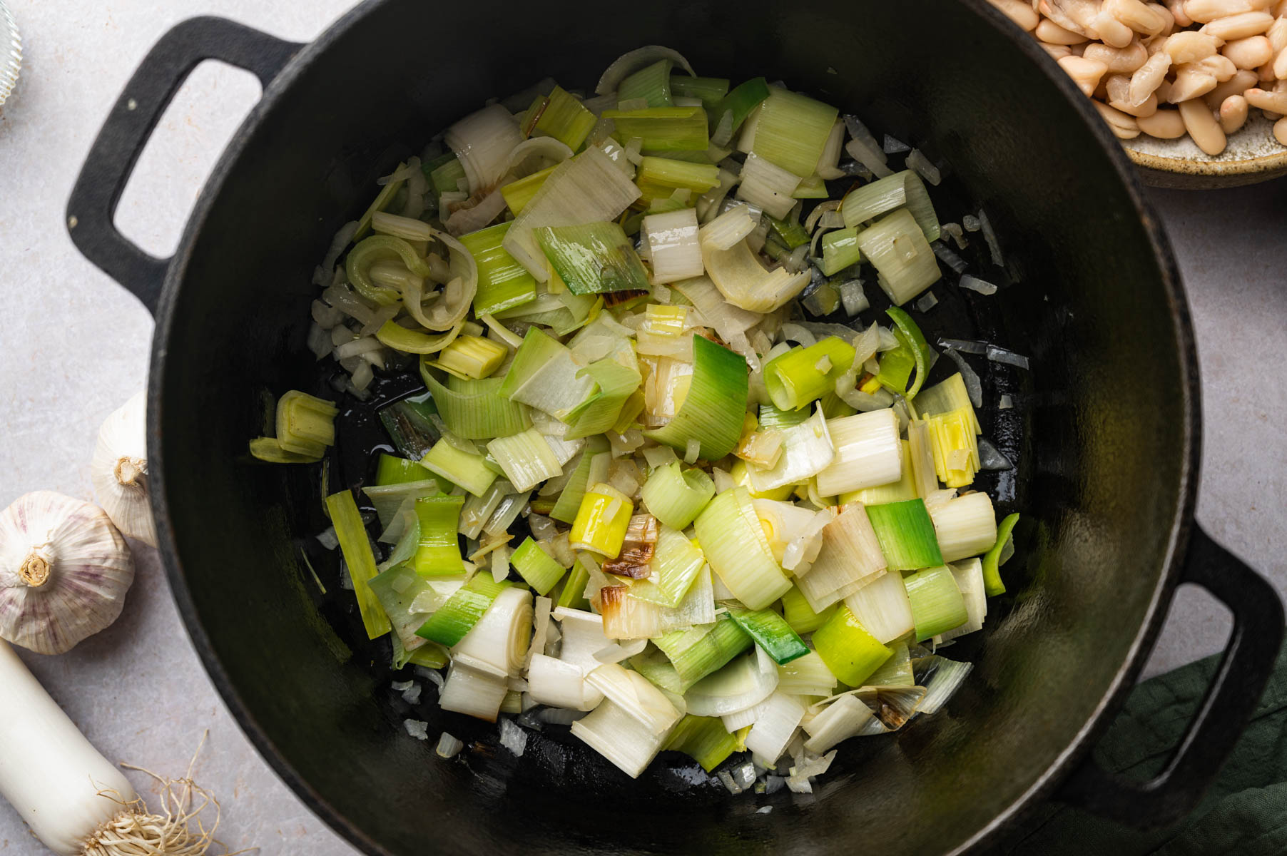 Chopped leeks sautéing in a black pot, next to garlic cloves and a bowl of white beans.