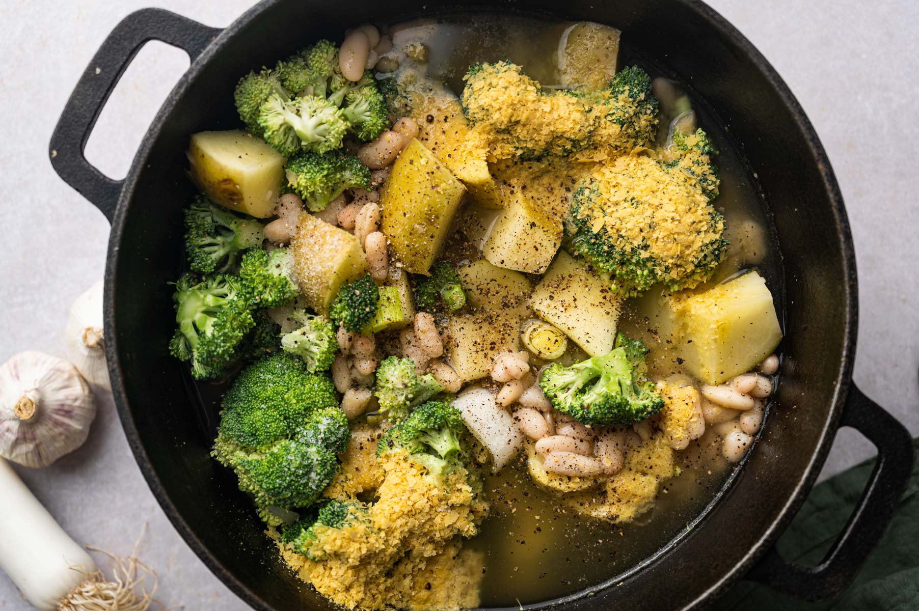 A pot containing broccoli, potatoes, white beans, and a sprinkle of seasoning in a clear broth. Garlic cloves and a leek are visible on the side.