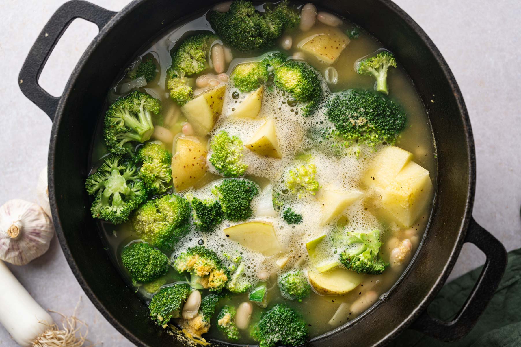 A black pot filled with broccoli, potato chunks, and beans in a broth. A garlic bulb and a leek are partially visible beside the pot.