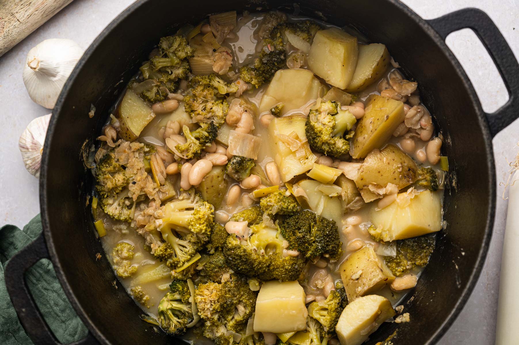 A pot of cooked mixed vegetables, including broccoli, potatoes, and white beans, in a light broth. Garlic cloves are nearby.