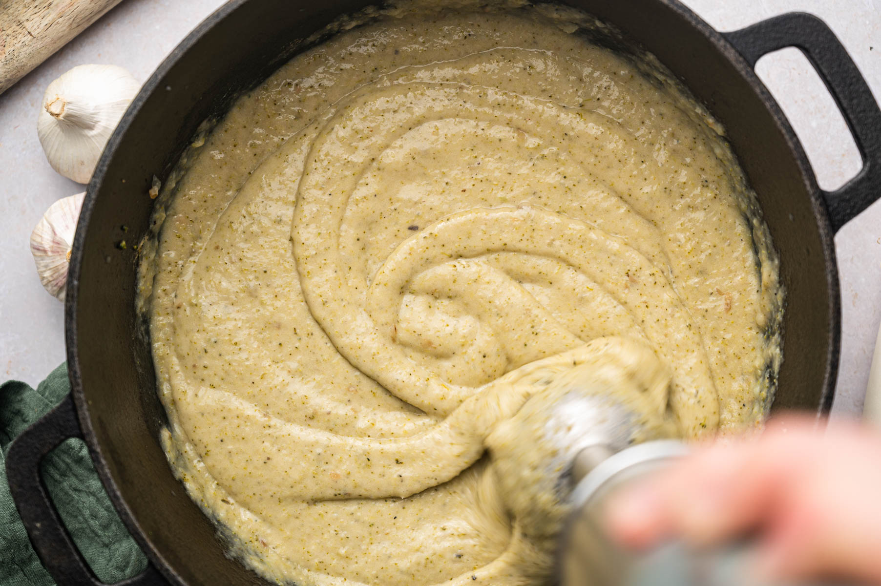 A hand blends a thick, yellow-green mixture in a black pot using an immersion blender. Garlic bulbs and a green cloth are partially visible in the background.