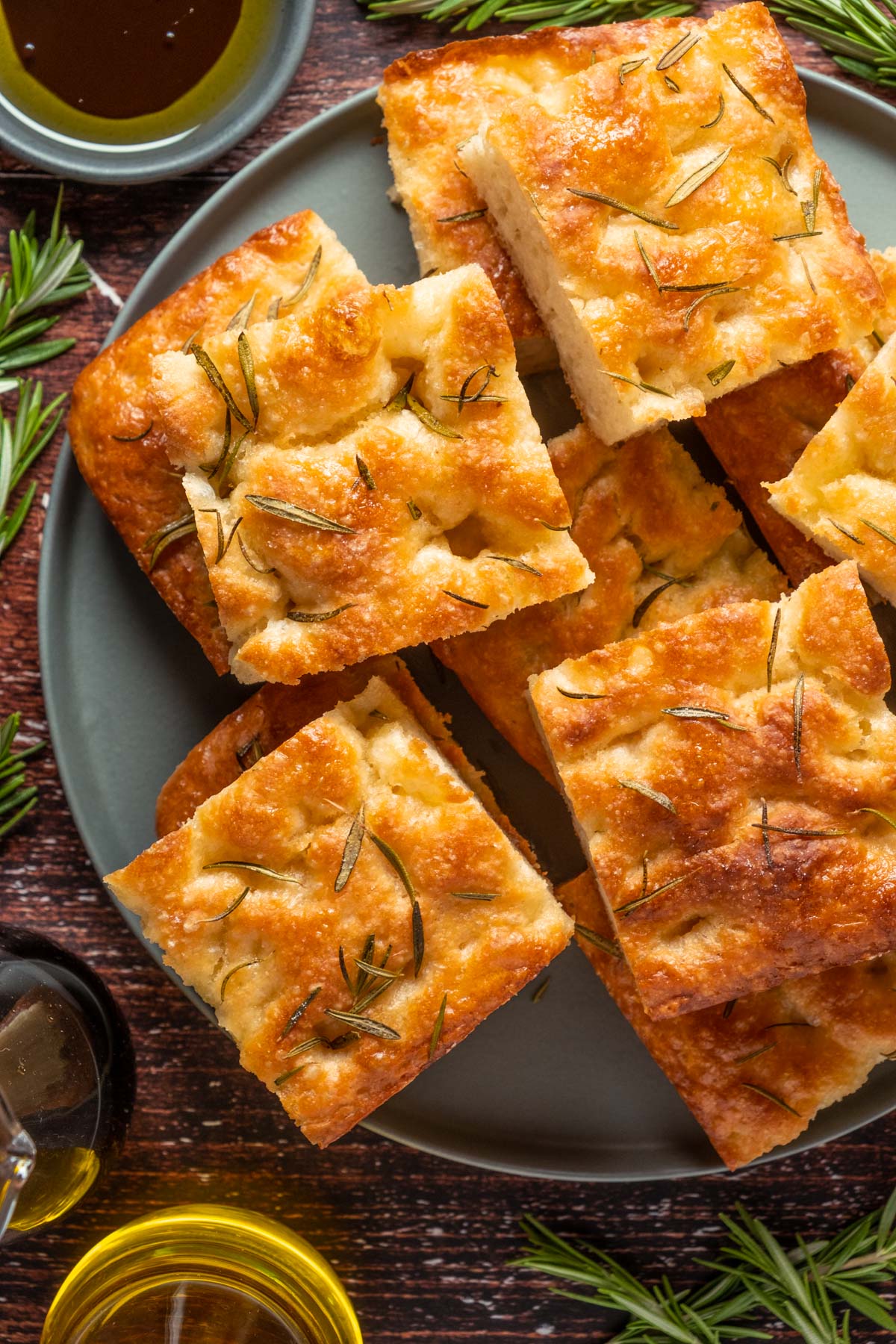 A plate of golden-brown focaccia slices garnished with rosemary, surrounded by sprigs of rosemary and small bowls of olive oil and balsamic vinegar, making for an appetizing setup alongside a steaming bowl of butternut squash soup.