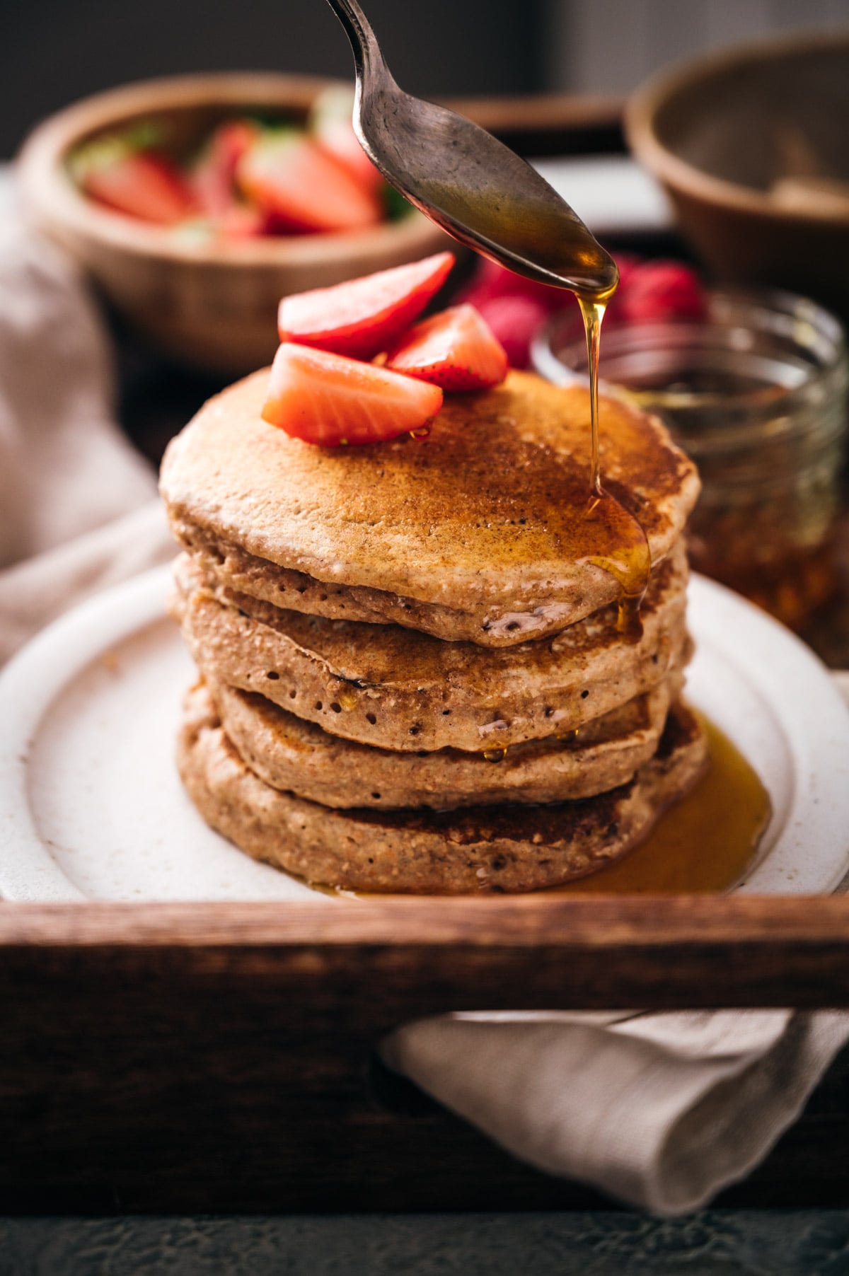 Stack of pancakes topped with sliced strawberries being drizzled with syrup on a white plate, with a bowl of strawberries in the background.