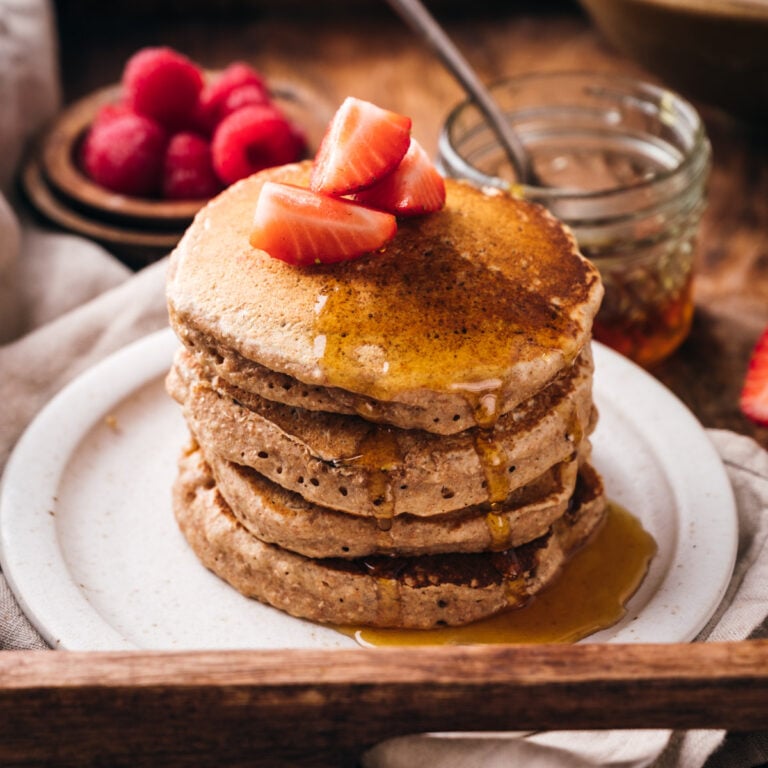 A stack of vegan pancakes topped with sliced strawberries and drizzled with syrup sits enticingly on a white plate, with a jar of syrup and a bowl of raspberries in the background.