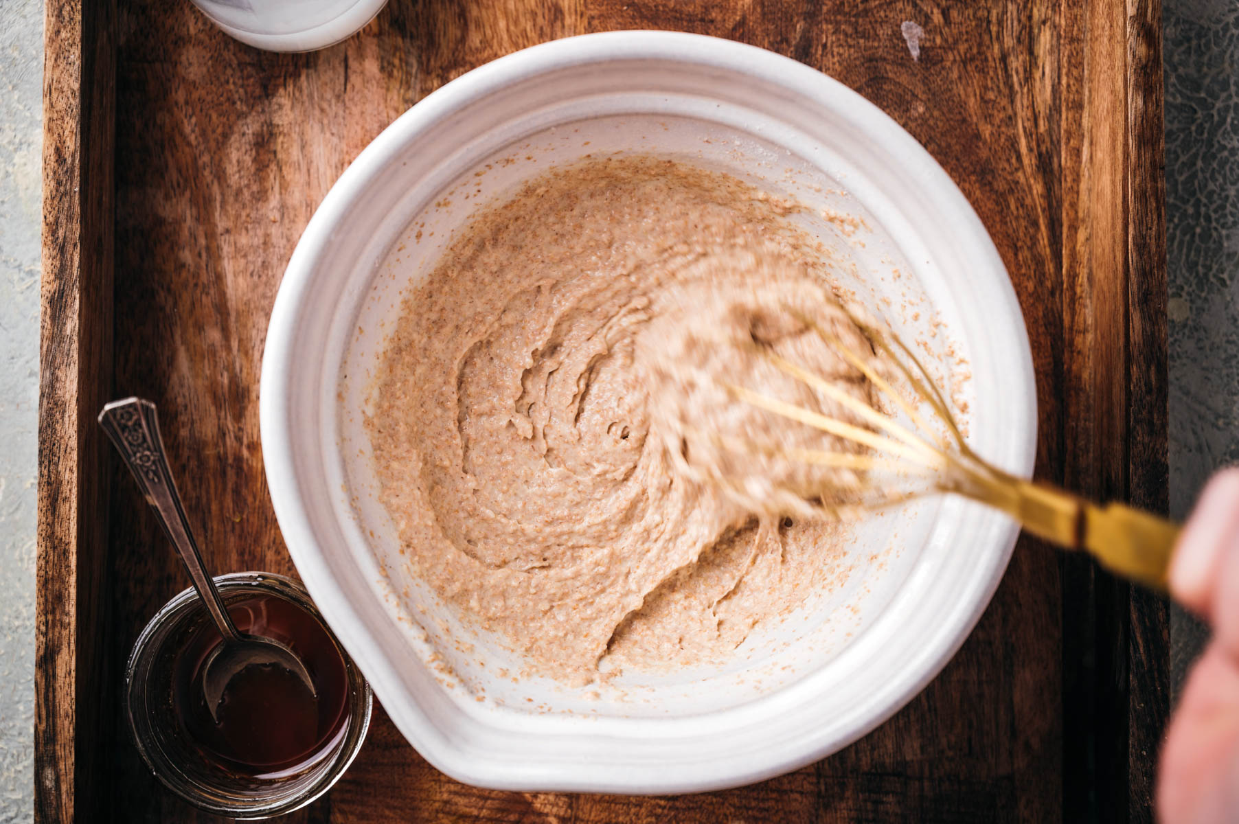 A bowl with a whisk is mixing a brown batter on a wooden surface. A small jar with syrup and a spoon is placed next to the bowl.