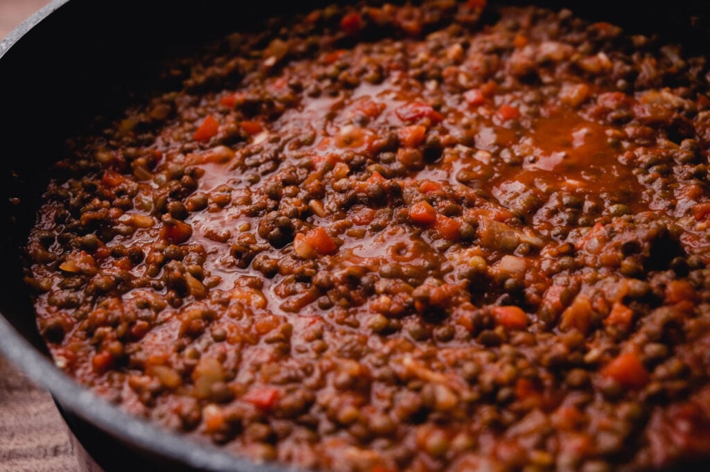 Close-up image of a skillet filled with cooked lentils and tomatoes, showing a textured surface of the simmering dish.
