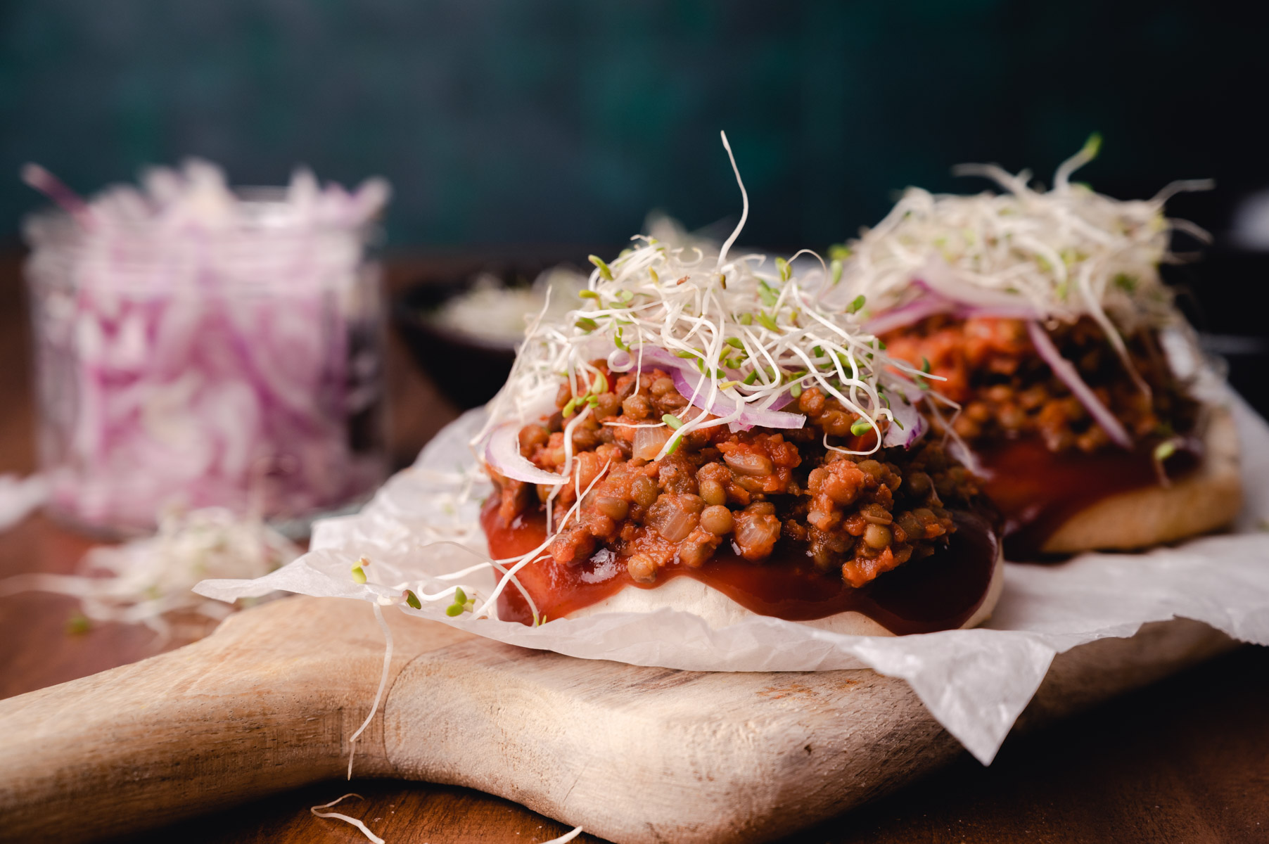 Two lentil sloppy joes topped with sprouts on a wooden cutting board, with a jar of pickled onions in the background.