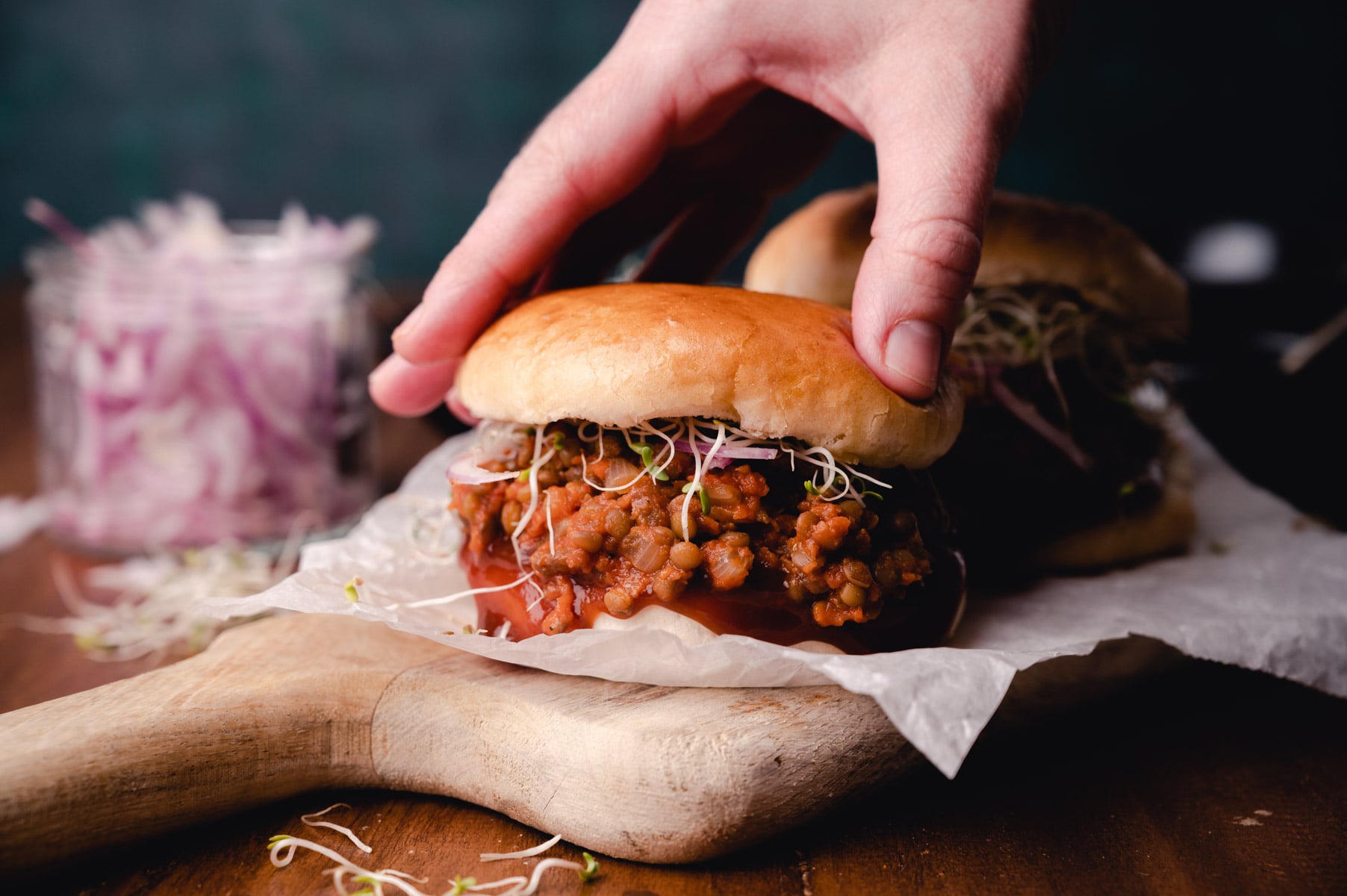 A hand placing the top bun on a lentil burger garnished with sprouts, served on a wooden board with onion slices in the background.