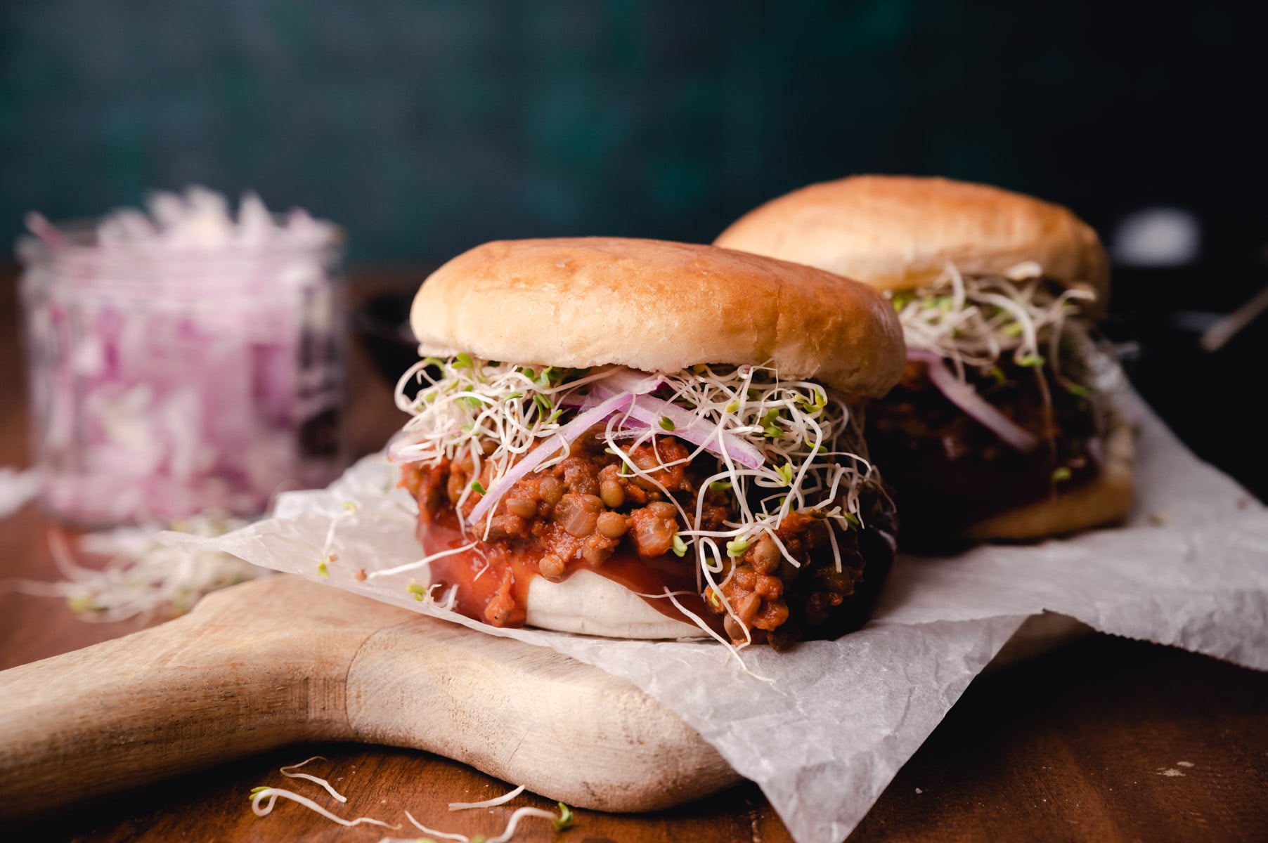 Two barbecue pulled pork sandwiches topped with sprouts on a wooden board, with a side of coleslaw in a glass container.