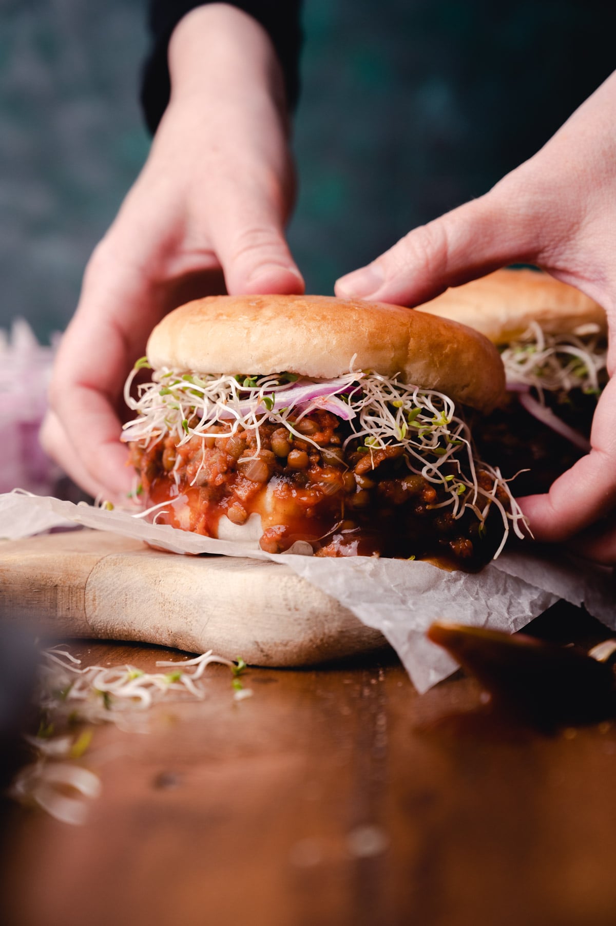 Hands holding a sloppy joes with sprouts and a bean patty on a wooden board.