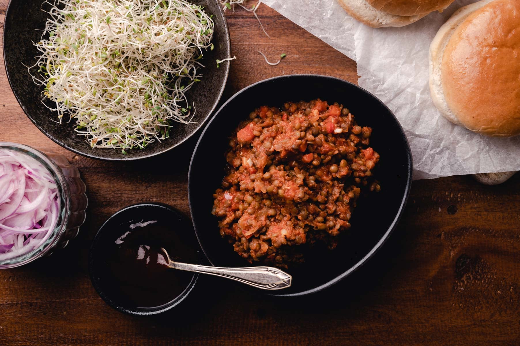 A bowl of cooked sloppy joes next to a bowl of sprouts, sliced onions, buns, and a small bowl of sauce, all arranged on a wooden table.