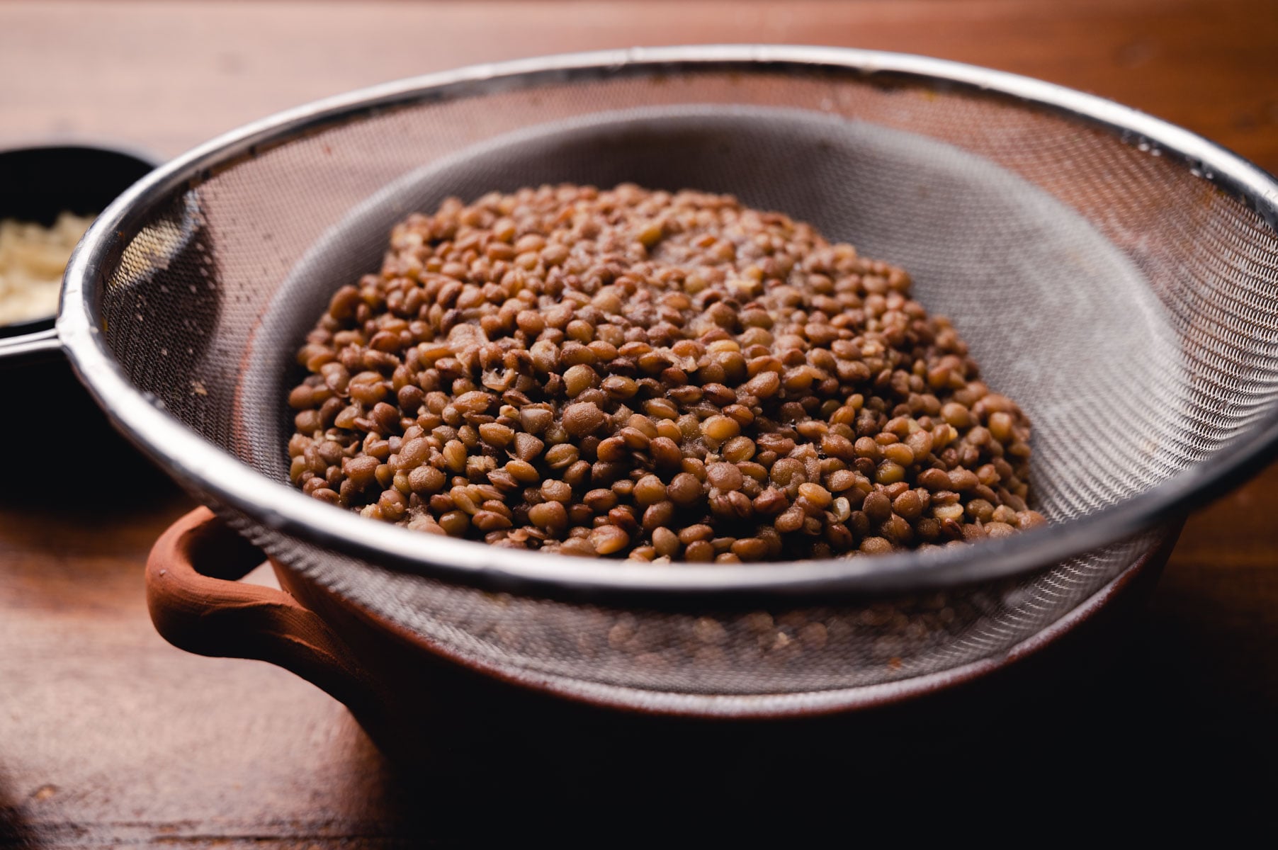 Cooked lentils in a metal sieve on a wooden table, with garlic cloves in the background.