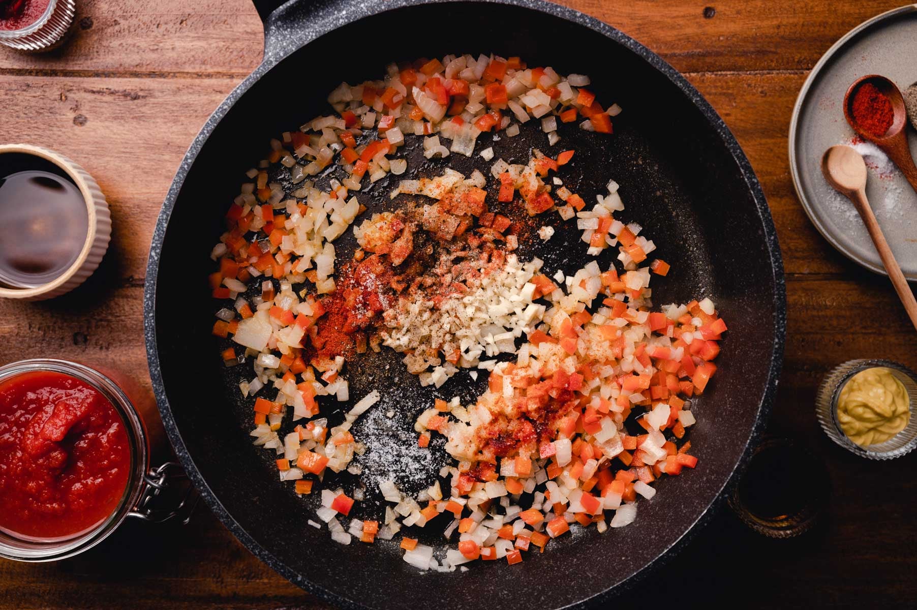 Diced onions, carrots, and celery sautéing with spices in a black cast iron skillet, with cooking utensils and sauce on the side.