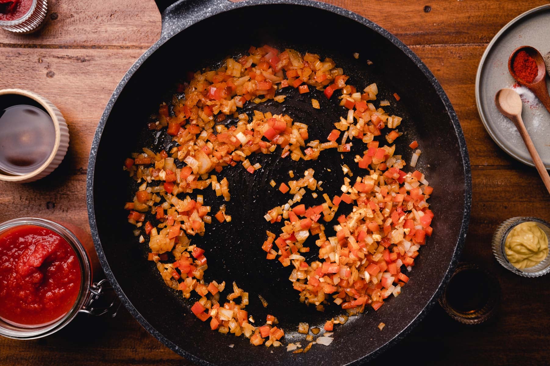 Diced onions and tomatoes sautéed in a black cast iron skillet, surrounded by spices and cooking utensils on a wooden table.