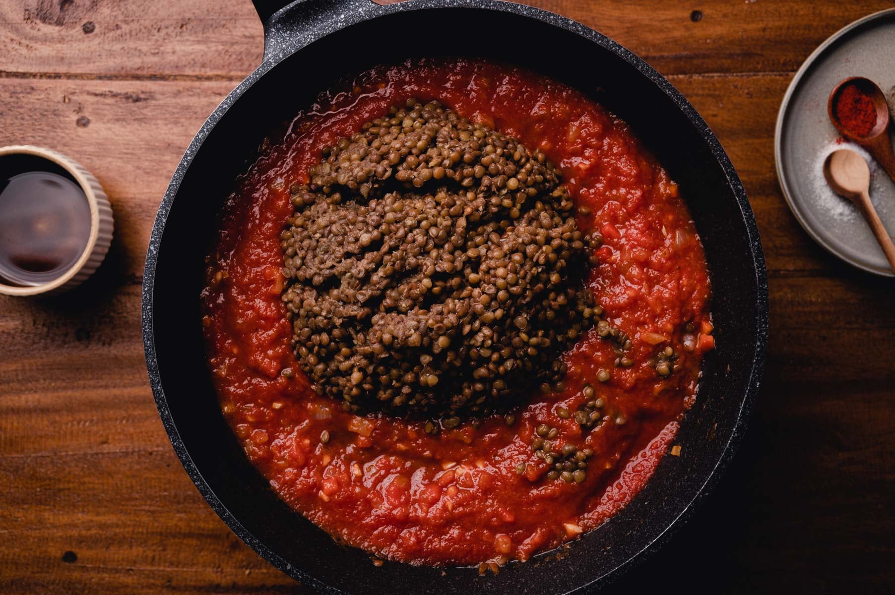 A skillet with a tomato sauce base topped with cooked lentils, photographed on a wooden surface alongside spices.