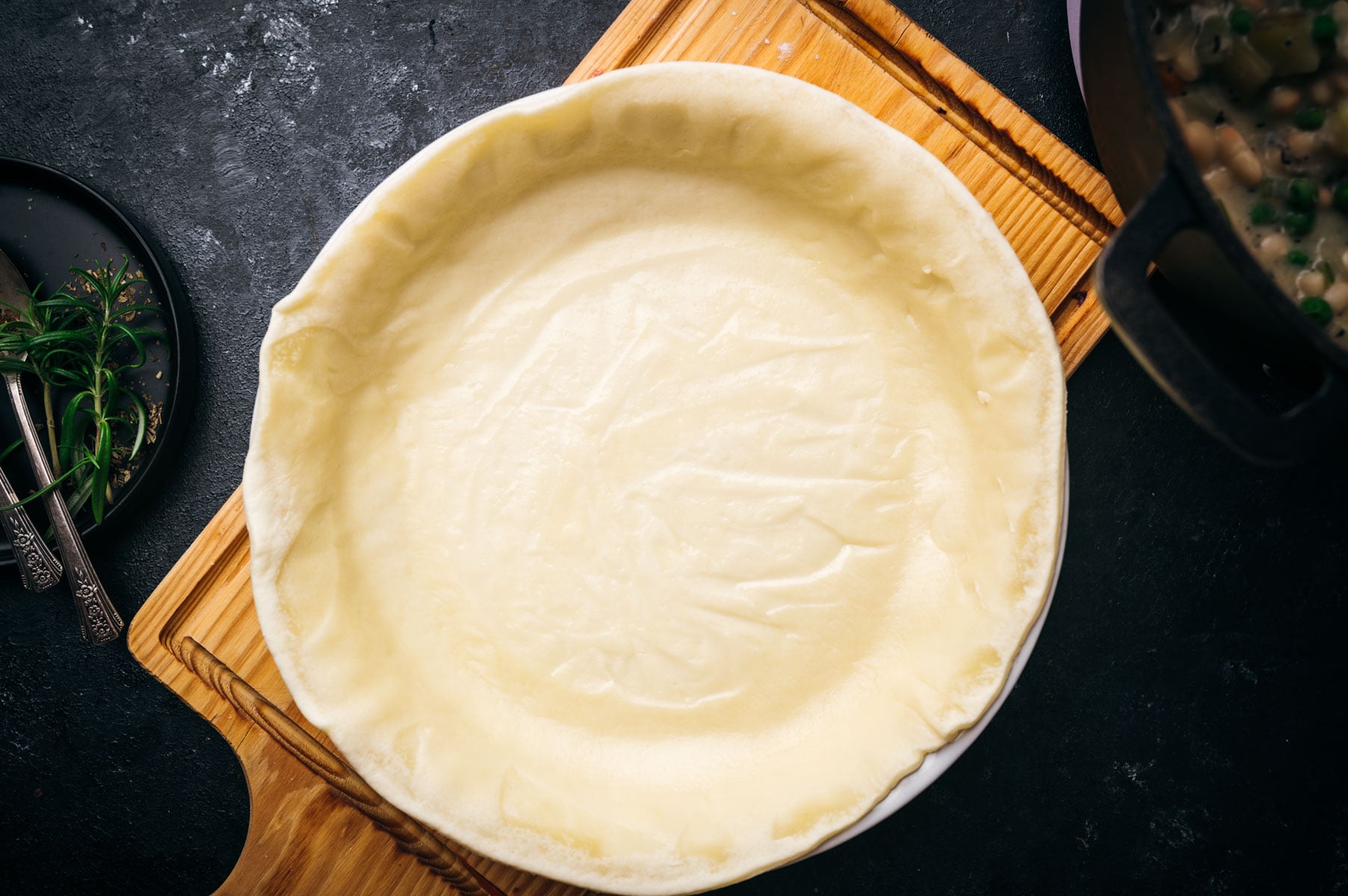 Unbaked pie crust in a dish on a wooden board, ready for filling, with rolling pin and herbs in background on a dark surface.
