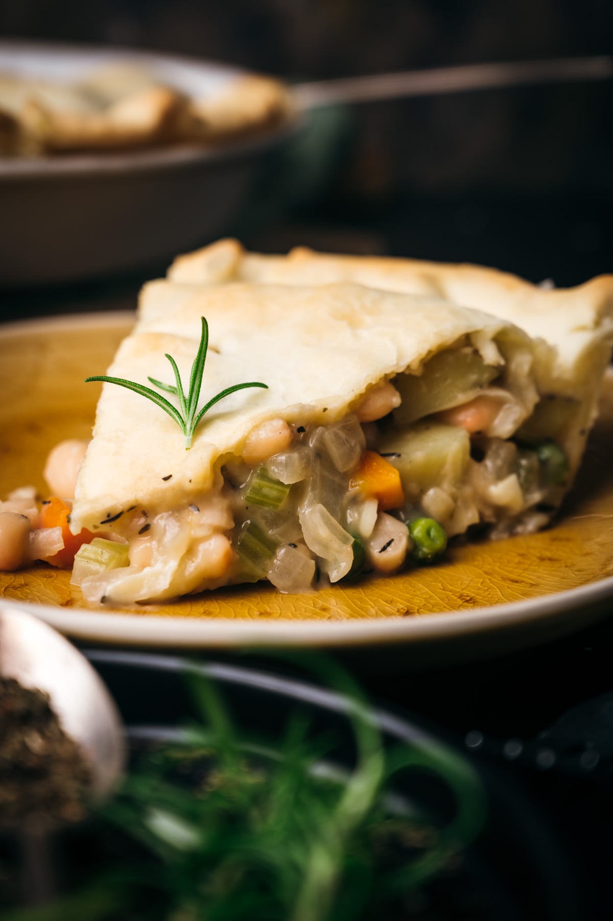 A slice of vegetable pot pie with a flaky crust, garnished with a sprig of rosemary, served on a wooden plate.