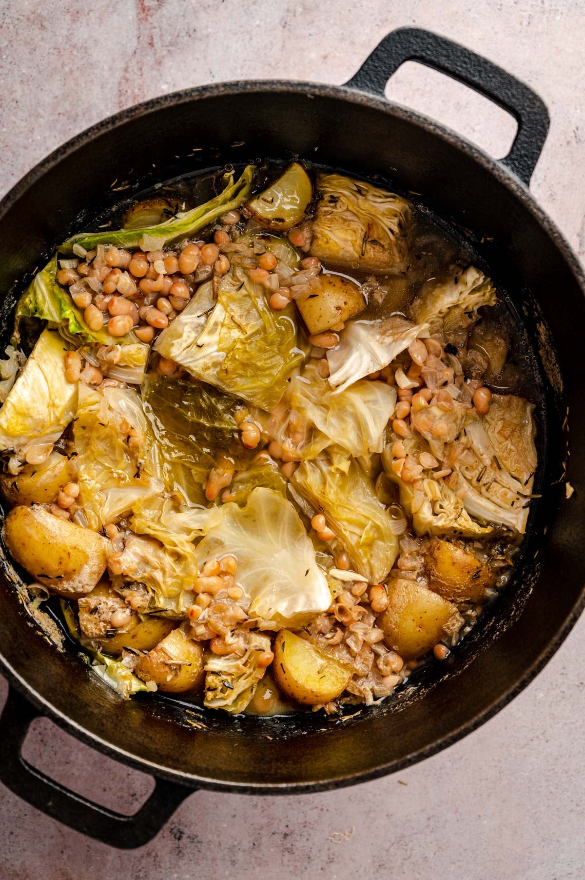 A cast iron pot filled with a cooked dish of cabbage, potatoes, and white beans in a savory broth.