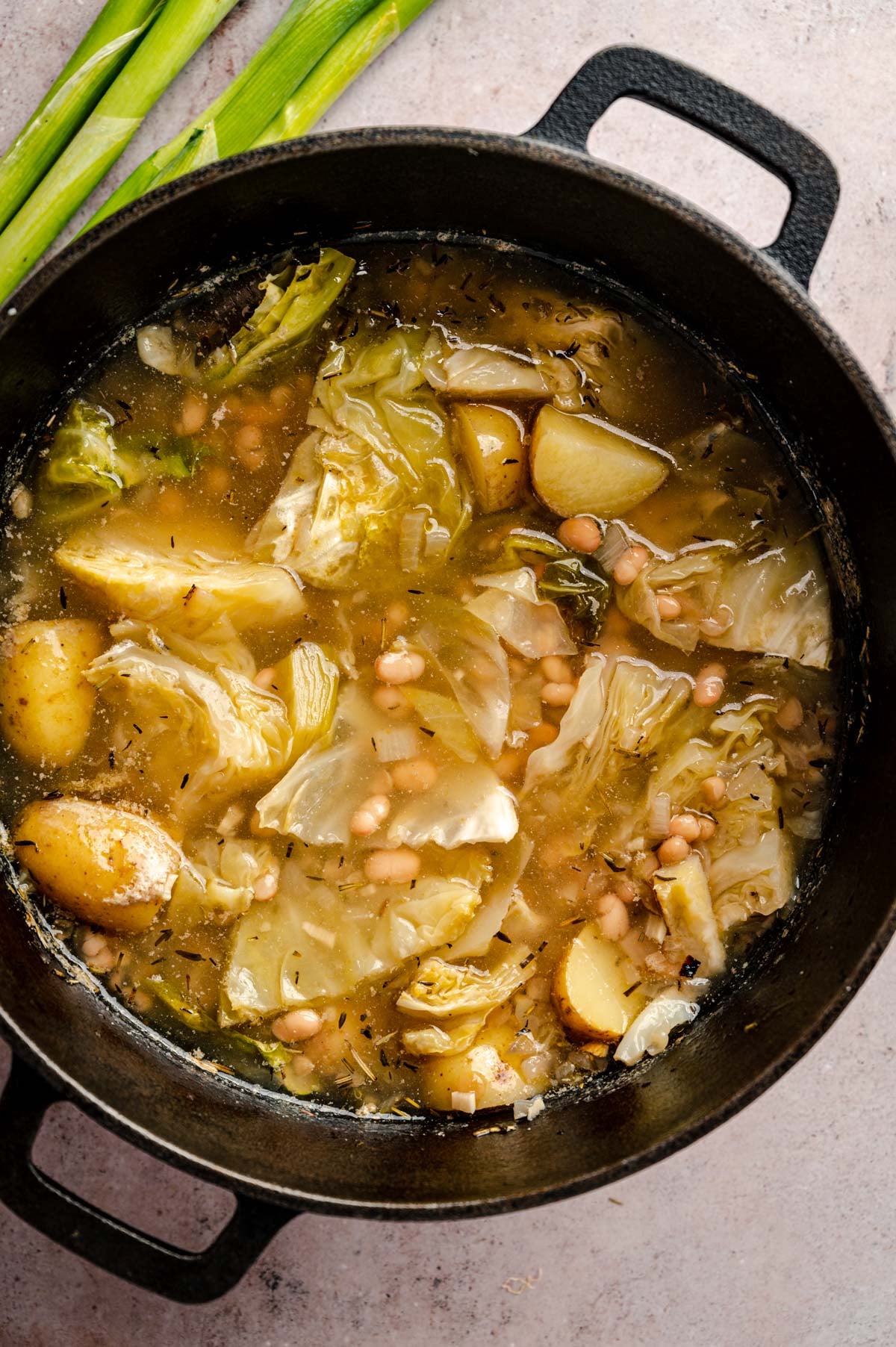 A pot of vegetable soup containing cabbage, potatoes, chickpeas, and broth. Green onion stalks are placed beside the pot.