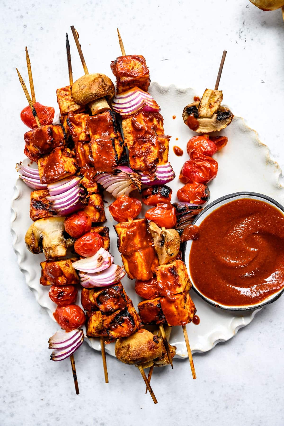 Grilled vegetable and tofu skewers with tomatoes, red onions, and mushrooms, served alongside a delicious pasta salad with a bowl of red dipping sauce on a white plate. Perfect if you're wondering what to serve for dinner!
