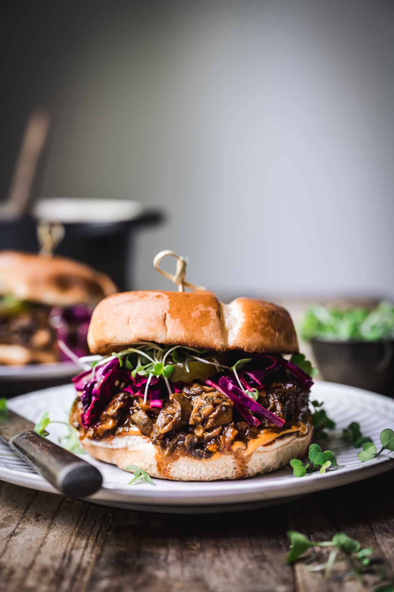 A sandwich with pulled meat, red cabbage slaw, and greens on a white plate, placed on a rustic wooden table. A butter knife is beside the plate. Serve with a side of pasta salad for a complete and delicious dish.