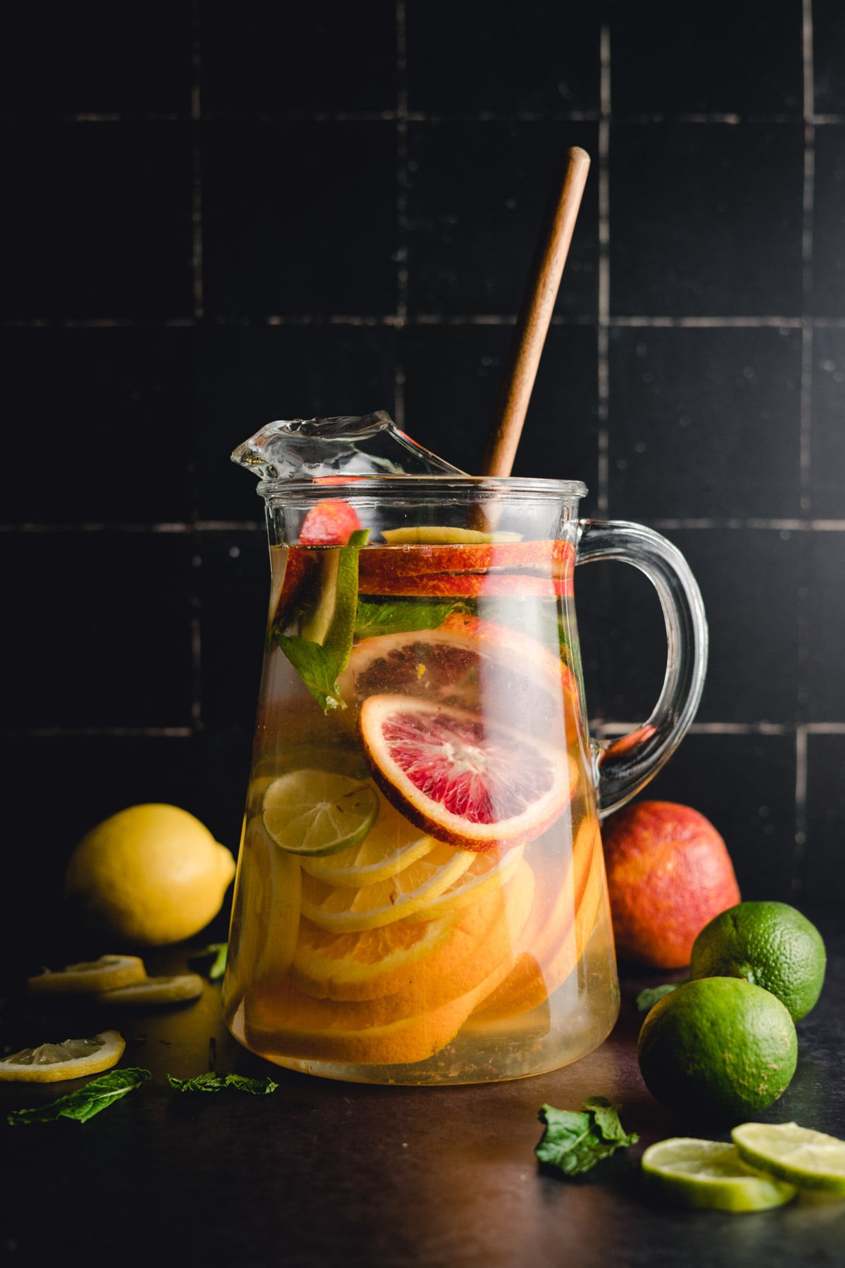 A clear glass pitcher filled with water, sliced citrus fruits including blood oranges, limes, and a lemon, with a wooden stirring stick. Whole fruits are scattered around the base.