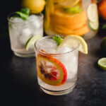 A close-up of a glass filled with ice cubes, garnished with a slice of lemon, a slice of orange, and a mint leaf. Another similar glass and a pitcher with lemon and lime slices are in the background.