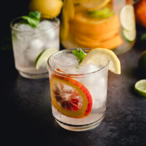A close-up of a glass filled with ice cubes, garnished with a slice of lemon, a slice of orange, and a mint leaf. Another similar glass and a pitcher with lemon and lime slices are in the background.