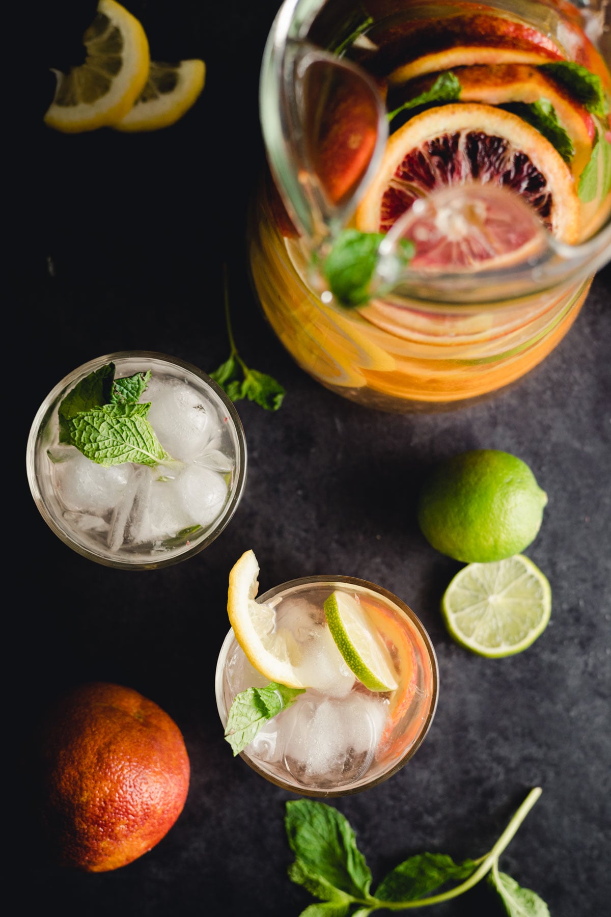 Overhead view of a pitcher filled with citrus-infused water surrounded by lemons, limes, and two glasses of iced citrus drink garnished with mint leaves.