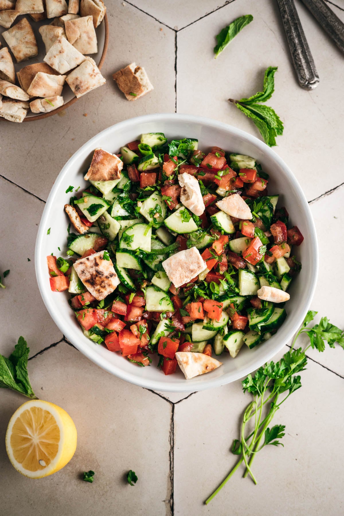 A bowl of fresh salad containing diced cucumbers, tomatoes, herbs, and pieces of pita bread on a tiled surface with a lemon and scattered herbs nearby.