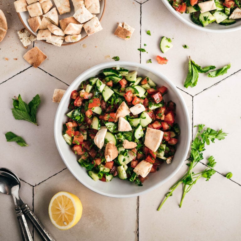 A white bowl filled with chopped cucumber, tomato, and herbs, garnished with pita bread pieces. Fresh mint, cilantro, a lemon half, and some scattered pita pieces are nearby, with spoons in the corner—perfect for a refreshing fattoush salad.