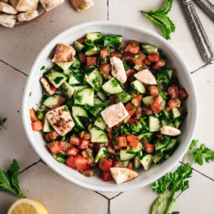 A bowl of chopped cucumber, tomato, parsley, and pita bread pieces on a tiled surface, reminiscent of a classic fattoush salad. Mint leaves, a lemon slice, and parsley sprigs are scattered around for a fresh touch.