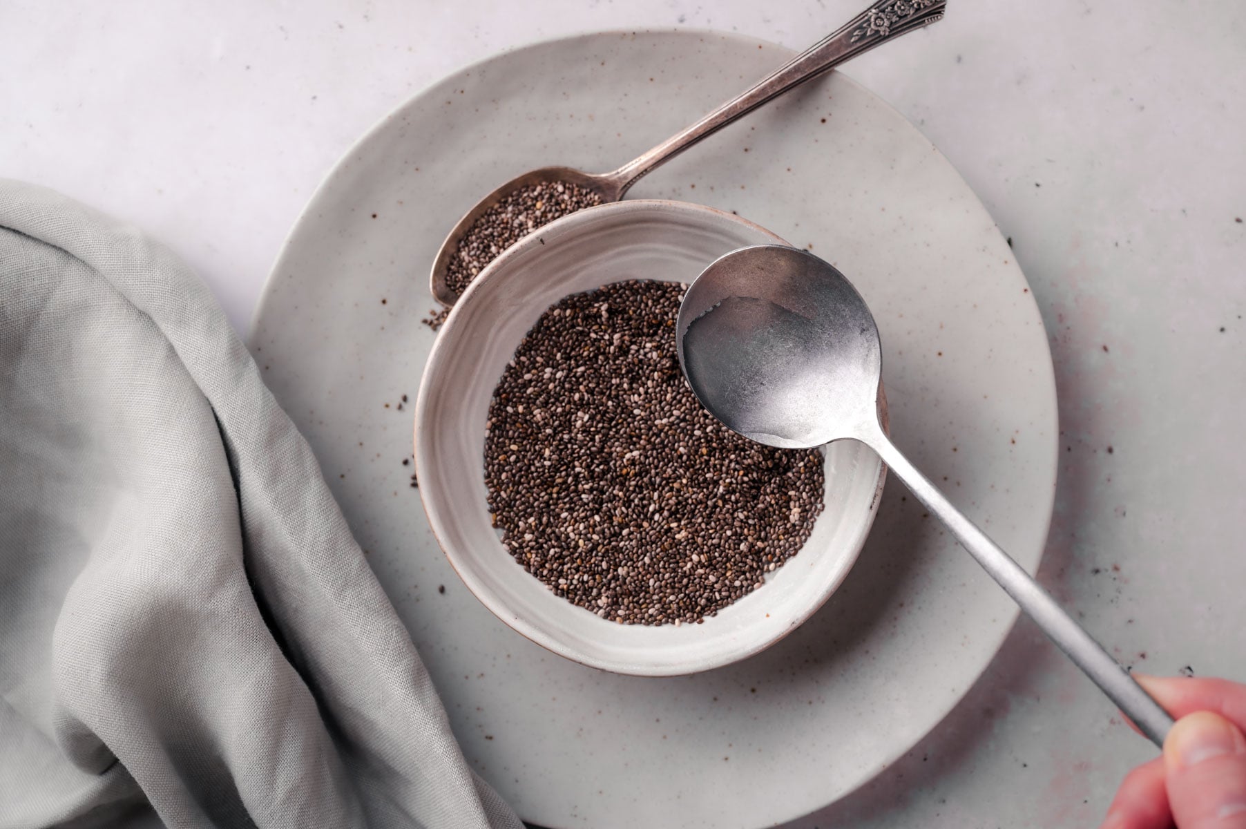 A bowl of chia seeds with a spoon inside and another spoon outside, placed on a plate with a gray cloth beside it on a light surface.