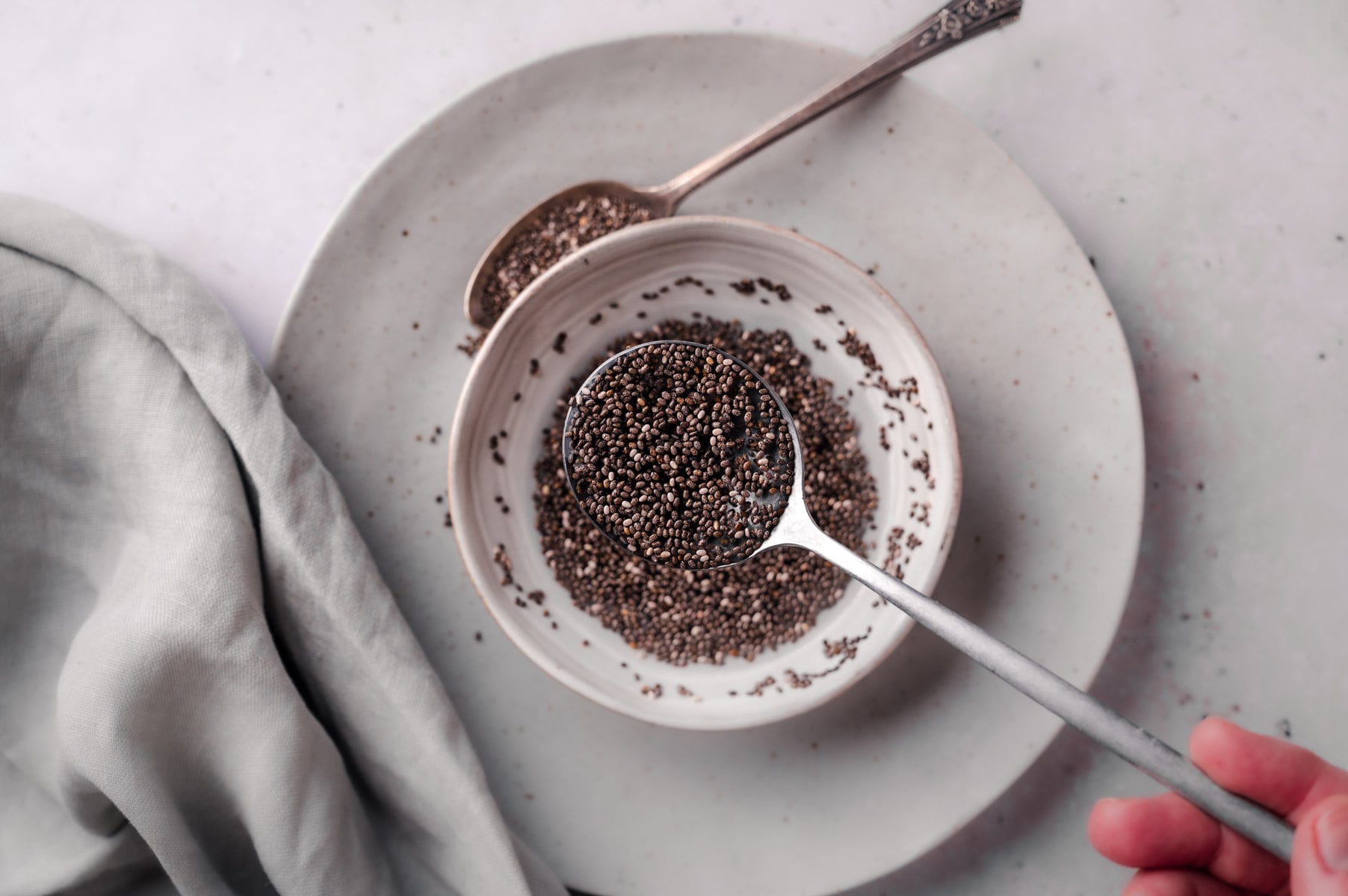 A silver spoon holding chia seeds is placed over a white bowl filled with more chia seeds. Another spoon and a light gray cloth are seen in the background on a plate.