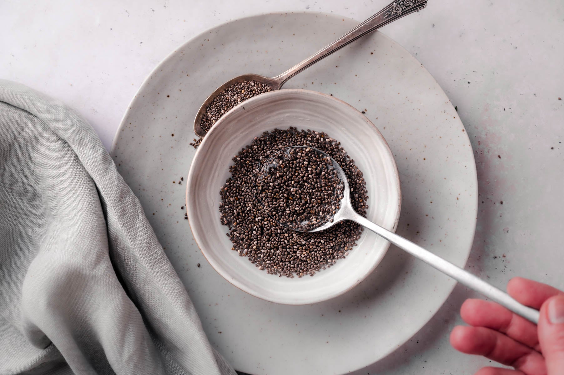 A hand holding a spoon over a bowl of chia seeds on a plate with two spoons, one inside the bowl and another on the plate. A light gray cloth is draped on the left side of the frame.
