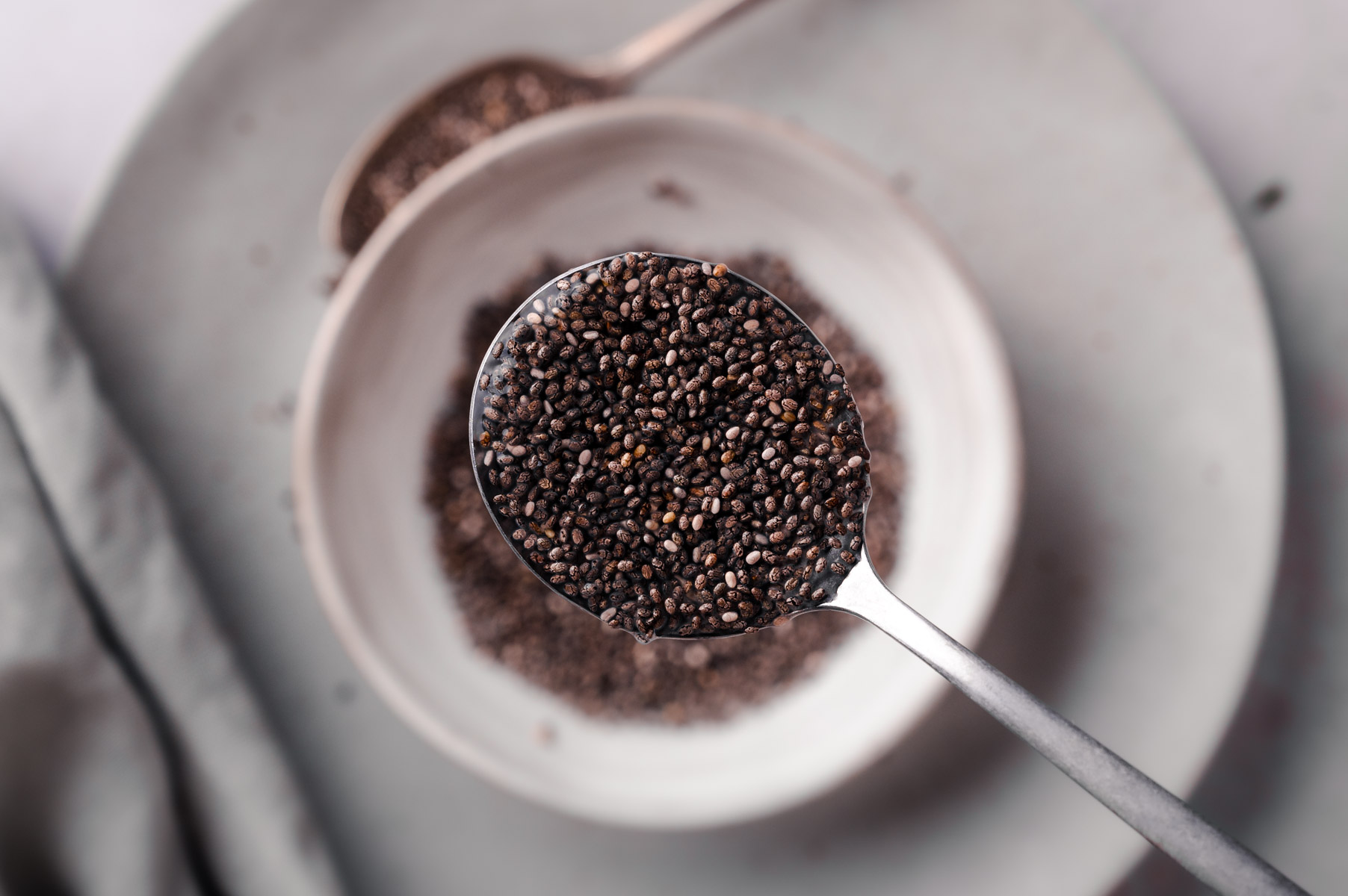 Close-up of a spoonful of chia seeds held over a white bowl filled with more chia seeds, with another small bowl of chia seeds in the background.