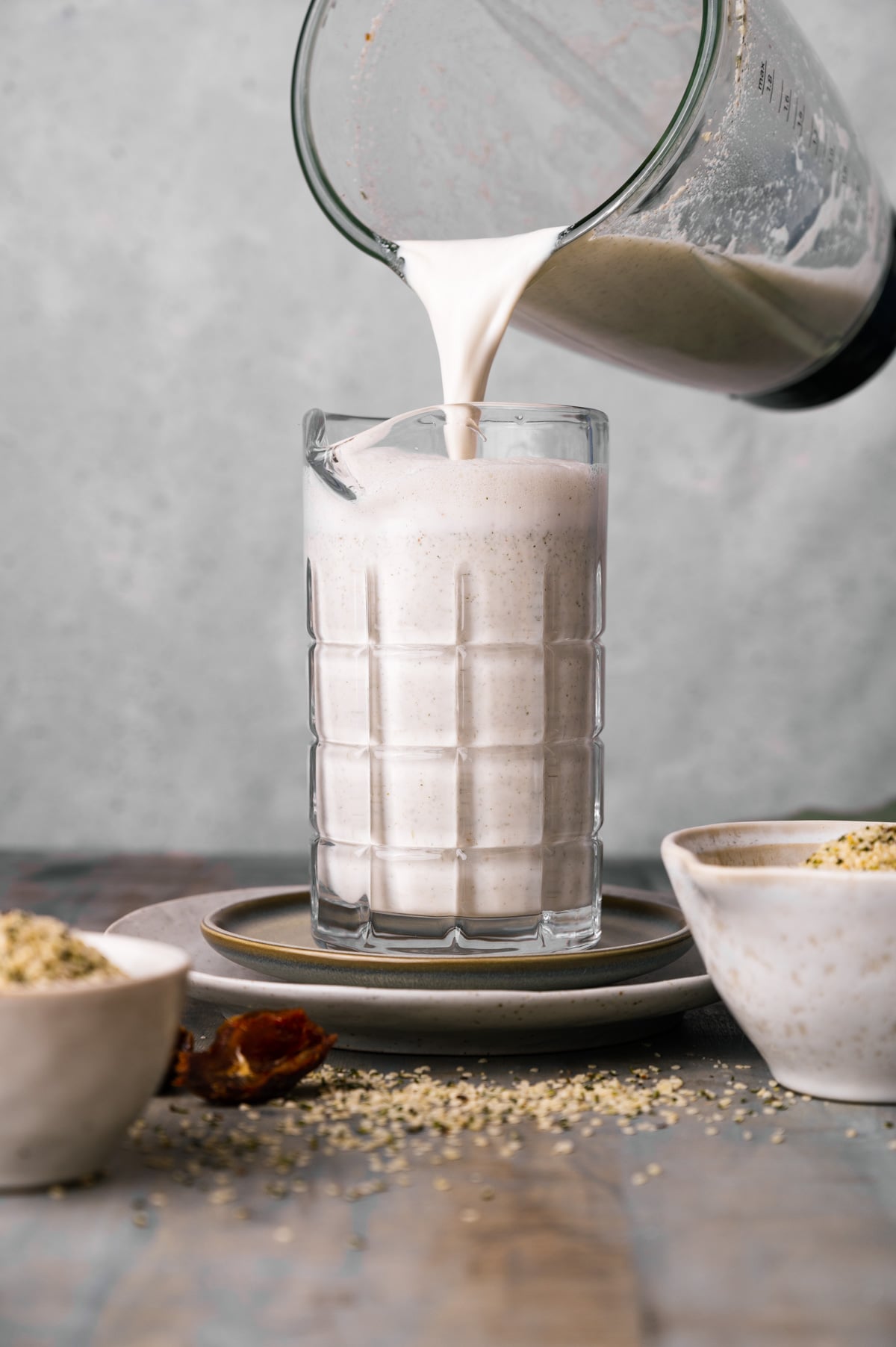 A blender pitcher pours a creamy hemp milk into a tall glass on a saucer. The table also holds two bowls with seeds and a date on a plate.