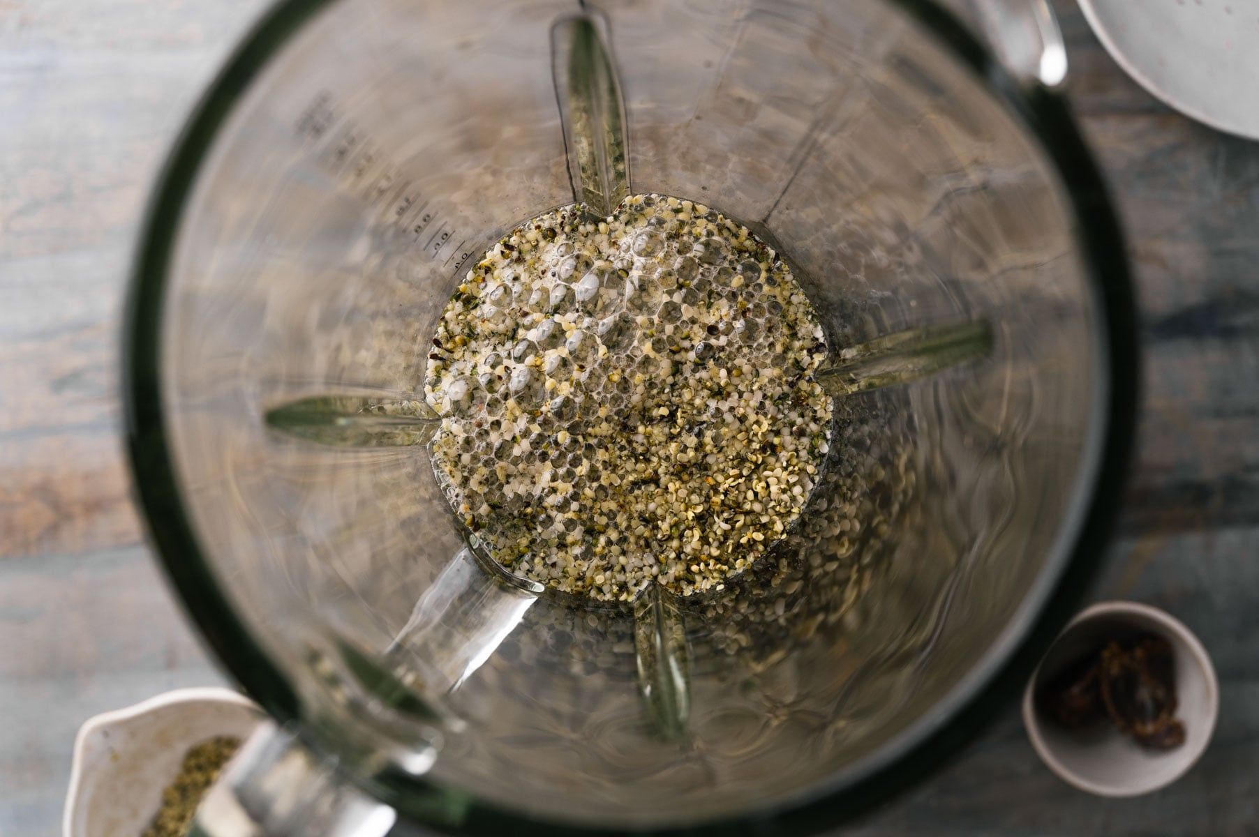 Top view of a blender with hemp seeds and liquid inside, sitting on a light-colored wooden surface. Nearby are two small bowls, one partially visible, containing additional ingredients.