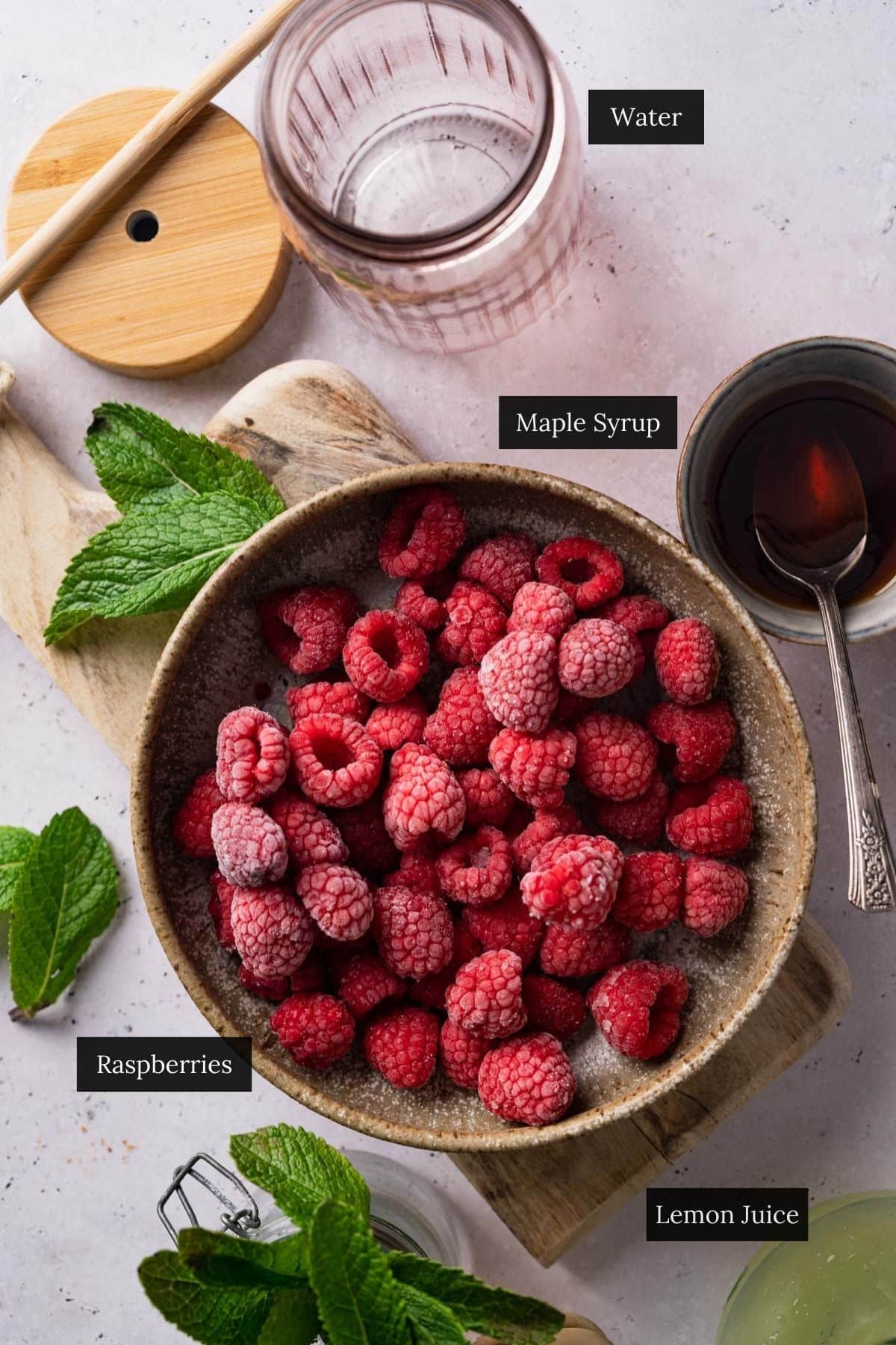 A bowl of raspberries on a wooden board with sprigs of mint, next to containers labeled water, maple syrup, and lemon juice.