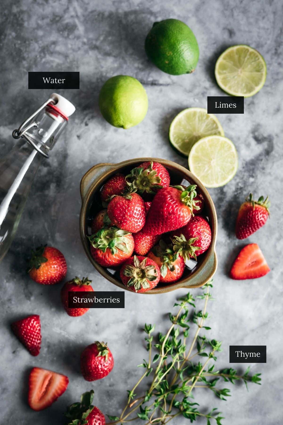 A bowl of strawberries surrounded by fresh limes, thyme, sliced strawberries, and a bottle of water on a gray countertop. Labeled ingredients include water, limes, strawberries, and thyme, creating the perfect setup for refreshing strawberry lime water.
