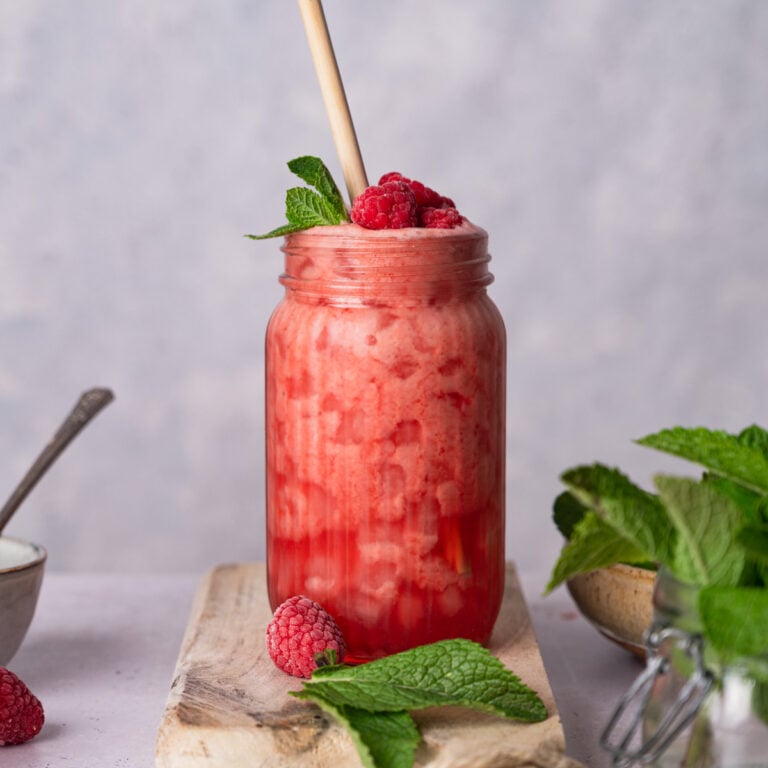 A glass jar filled with a pink lemonade, topped with raspberries and mint leaves, with a wooden straw. The jar is placed on a wooden board with additional mint leaves and a raspberry nearby.