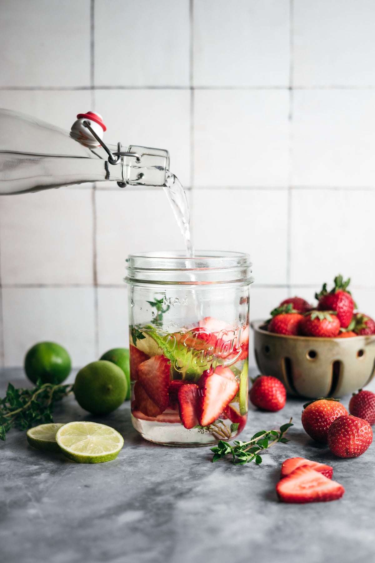 A glass bottle pours water into a jar filled with sliced strawberries, cucumbers, and green herbs. Whole strawberries, limes, and a bowl of strawberries are also on the table.