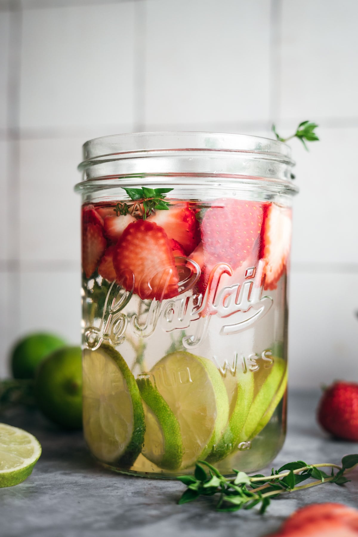 A mason jar filled with sliced strawberries and lime in liquid, garnished with herbs, in front of a tiled background.