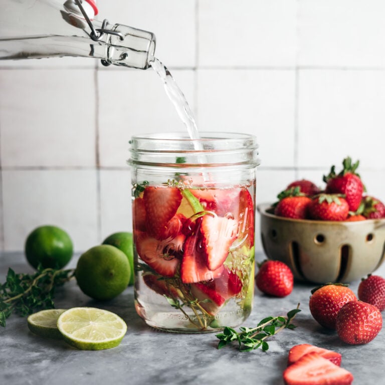 Water is being poured from a bottle into a mason jar containing sliced strawberries, lime, and herbs. Fresh limes and a bowl of strawberries are on the countertop.