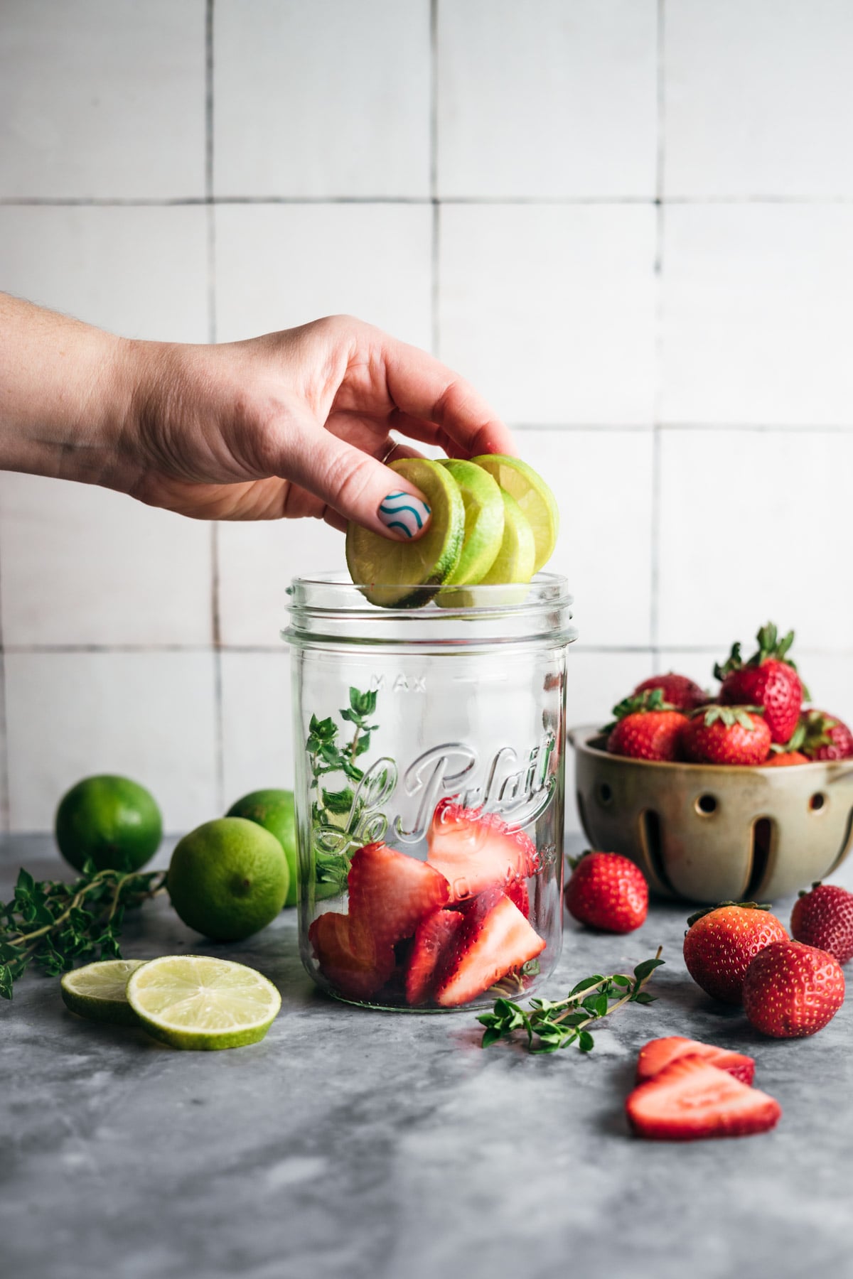 A hand places green apple slices into a mason jar filled with strawberries, surrounded by limes, cut fruit, and a bowl of strawberries on a countertop.